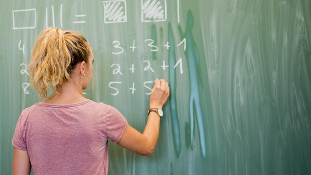 Das Bild zeigt eine Lehrerin von hinten. Sie steht an einer Schul-Tafel und schreibt darauf. Auf der Tafel steht eine mathematische Rechnung.