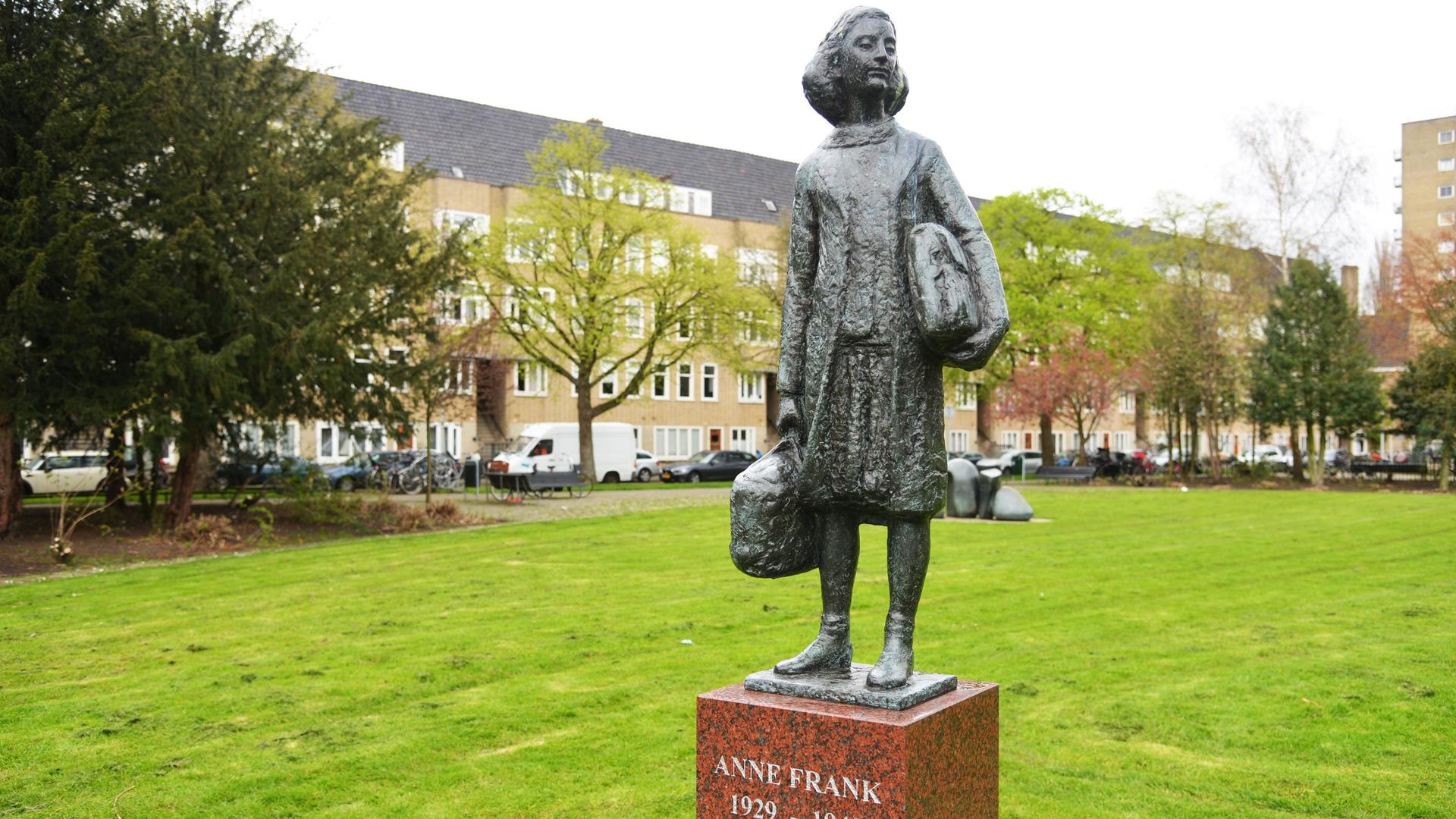 Ein Denkmal, das Anne Frank zeigt steht auf einer Wiese. Im Hintergrund sind Wohnhäuser. Auf dem Sockel des Denkmals steht "Anne Frank 1929 - 1945". Es zeigt Anne Frank mit einer Tasche in der rechten Hand und einer dicken Mappe unter dem linken Arm.