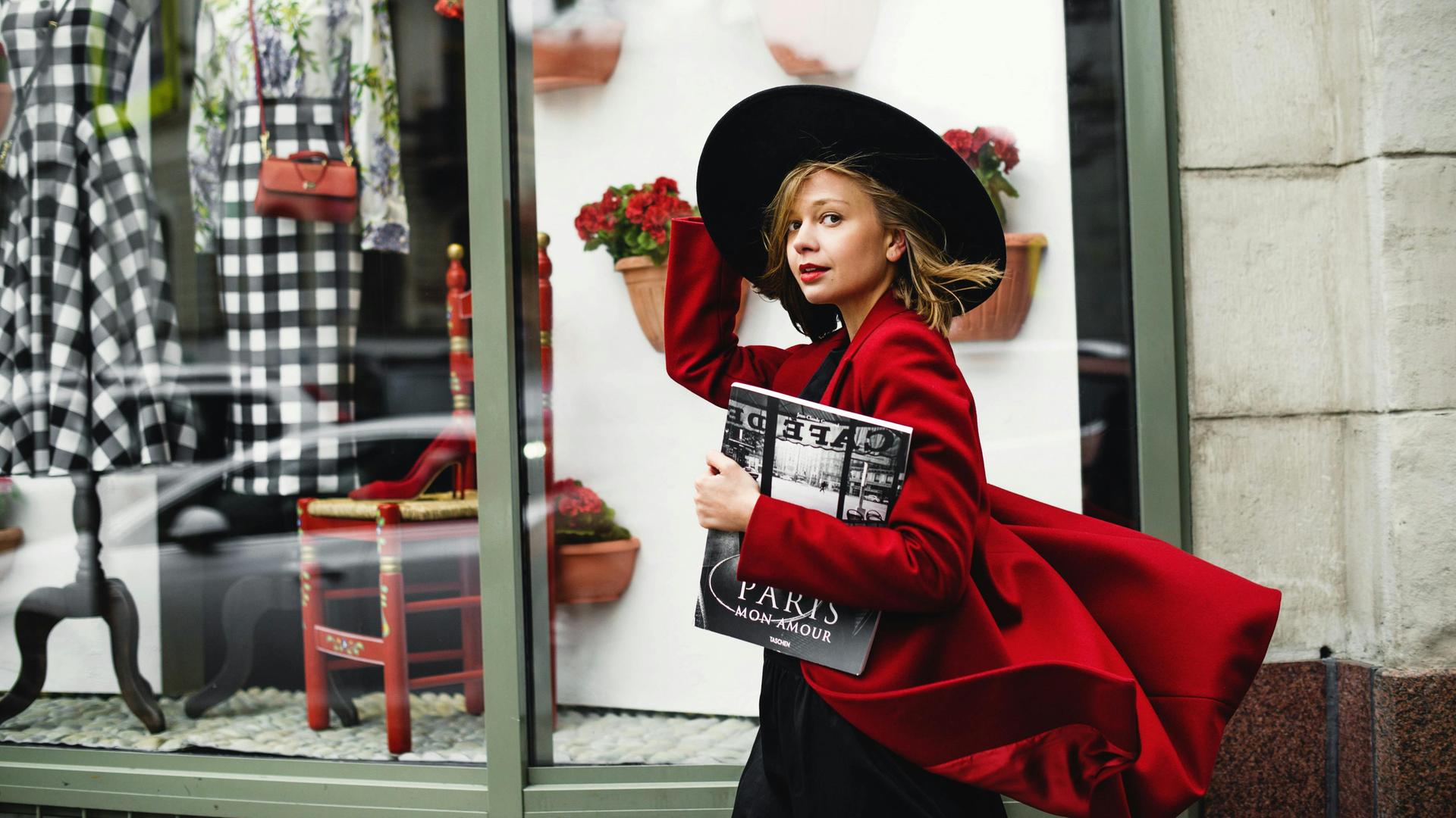 Eine junge Frau mit großem, schwarzen Hut und wehendem, roten Mantel geht an einem Pariser Geschäft vorbei und schaut dabei in die Kamera.