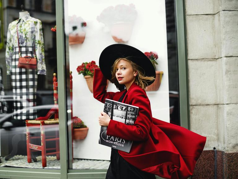 Eine junge Frau mit großem, schwarzen Hut und wehendem, roten Mantel geht an einem Pariser Geschäft vorbei und schaut dabei in die Kamera.
