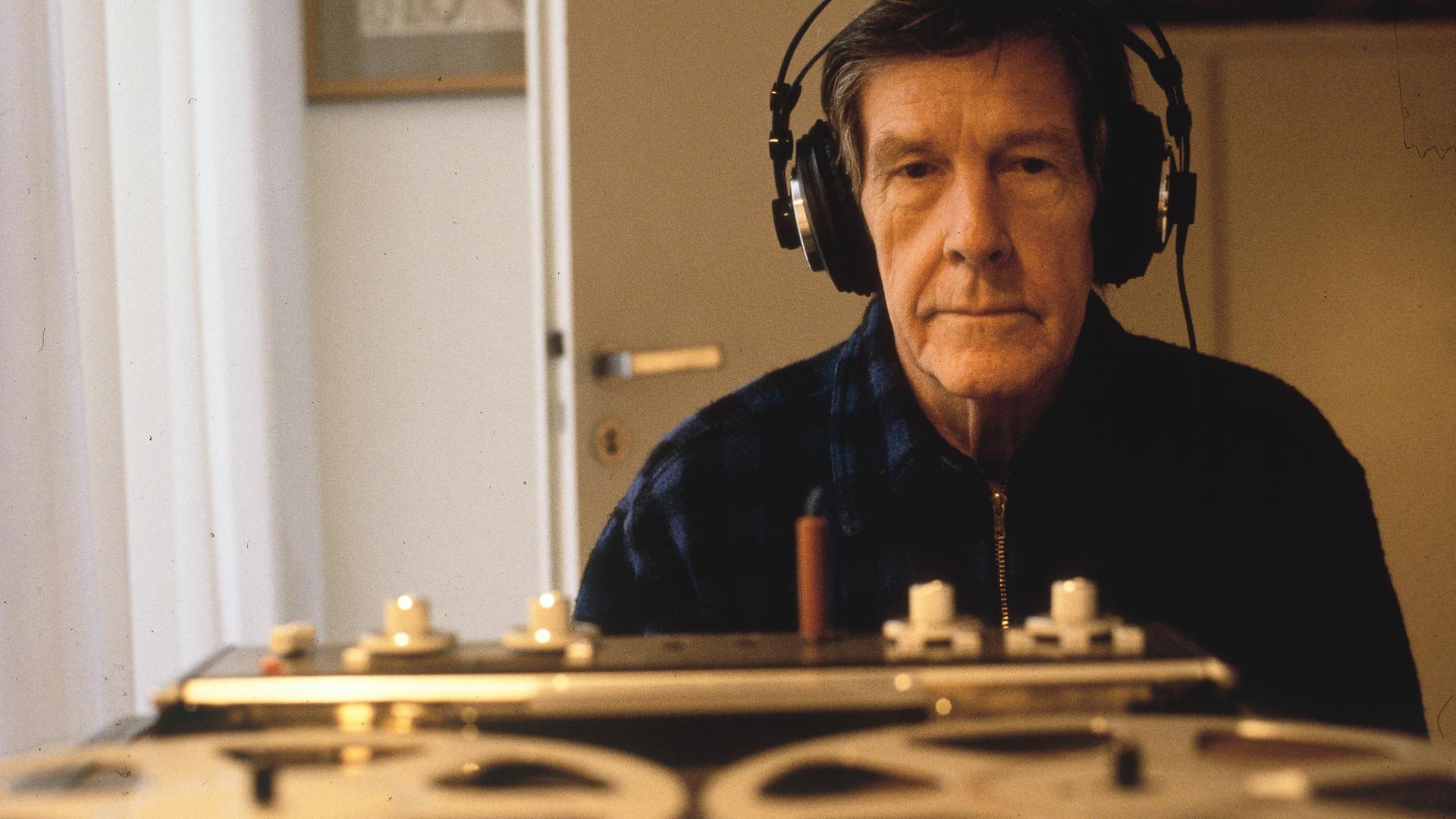 John Cage sitzt auf einem Foto aus dem Jahr 1981 mit Kopfhörern vor einem großformatigen Tonbandgerät in einem privaten Zimmer und hört konzentriert zu.