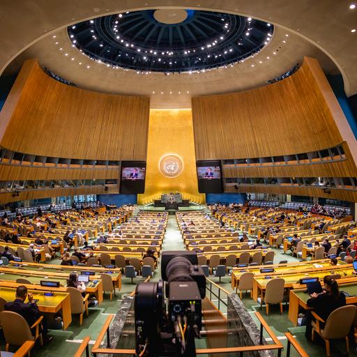 Das Bild zeigt den Sitzungssaal der Generalversammlung der Vereinten Nationen. Im Vordergrund sind viele Sitzreihen zu sehen. Im Hintergrund ist ein Redepult zu erkennen.
