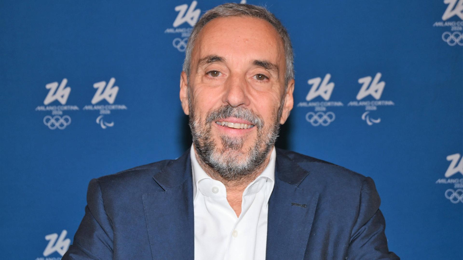 Andrea Varnier, CEO des Organisationskomitees der Olympischen Winterspiele 2026 in Mailand und Cortina d'Ampezzo; er sitzt vor einer blauen Wand mit dem olympischen Logo, trägt ein blaues Jackett, ein weißes Hemd und lächelt in die Kamera.