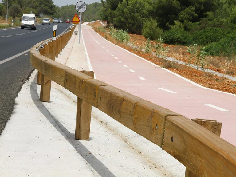 Eine Straße mit Pkws in Spanien. Straße und Radweg sind durch hölzerne Leitplanken getrennt.