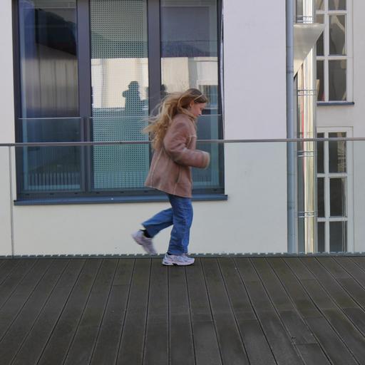 Ein Kind mit langem blonden Haar, Jeans, Jacke und Turnschuhen auf einer Dachterasse.
