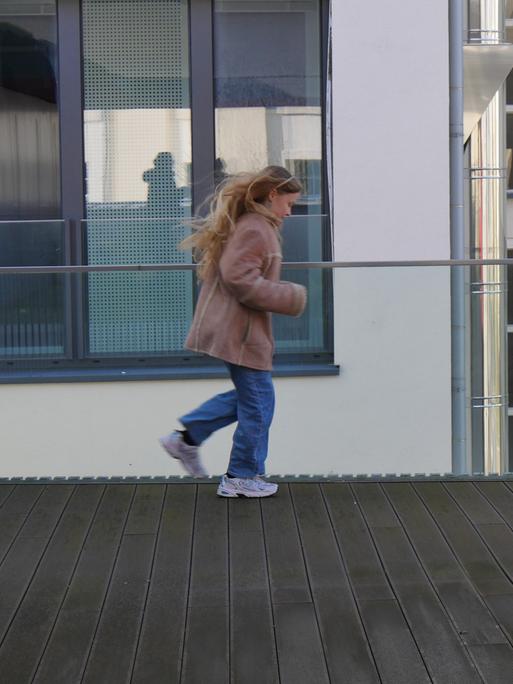 Ein Kind mit langem blonden Haar, Jeans, Jacke und Turnschuhen auf einer Dachterasse.