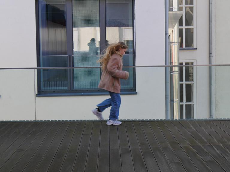 Ein Kind mit langem blonden Haar, Jeans, Jacke und Turnschuhen auf einer Dachterasse.