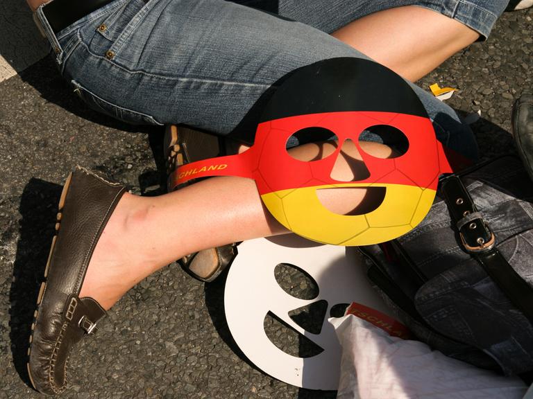 Public Viewing bei der WM 2006: Eine schwarz-rot-goldene Gesichtsmaske liegt auf dem Knie eines Teilnehmers, der auf dem Boden sitzt.