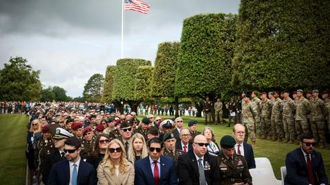 Zahlreiche Menschen laufen in einer Gedenkzeremonie über einen Friedhof. Im Hintergrund flattert eine amerikanische Flagge, an der Seite stehen einige Soldaten.