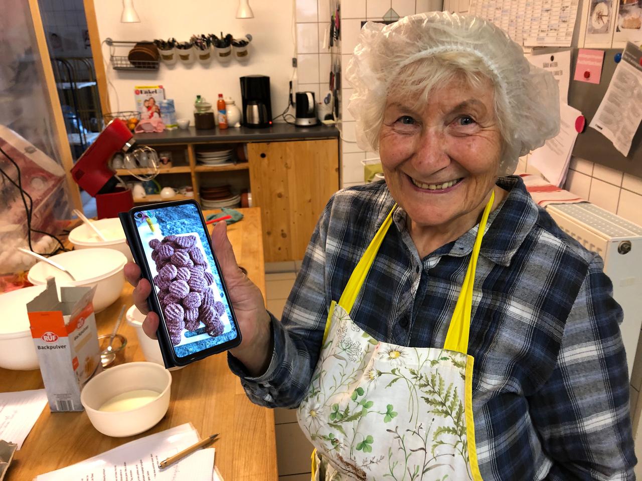 Oma Rosemarie steht mit einem Haarnetz in der Küche und lacht in die Kamera. Auf dem Handy zeigt sie ein Foto ihrer Lieblingskekse. Zu sehen sind viele braune, runde Kekse.
