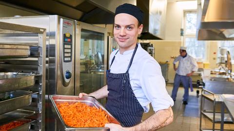Ein Koch, Patrick Wodni, präsentiert eine große Auflaufform mit Nudeln. Er steht in einer Großküche.