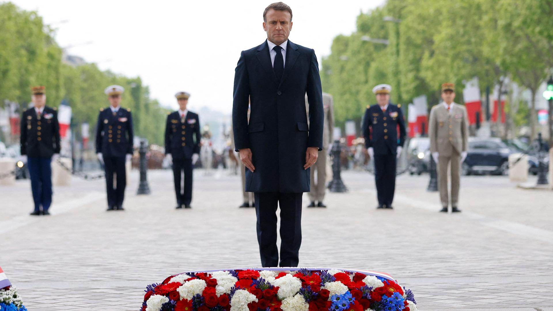 Frankreichs Präsident Macron steht mit ernstem Gesicht vor einem großen Blumenkranz.