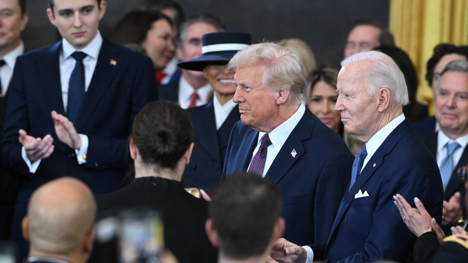 US-Präsident Biden und der künftige Präsident Trump sind umringt von mehreren Personen. 