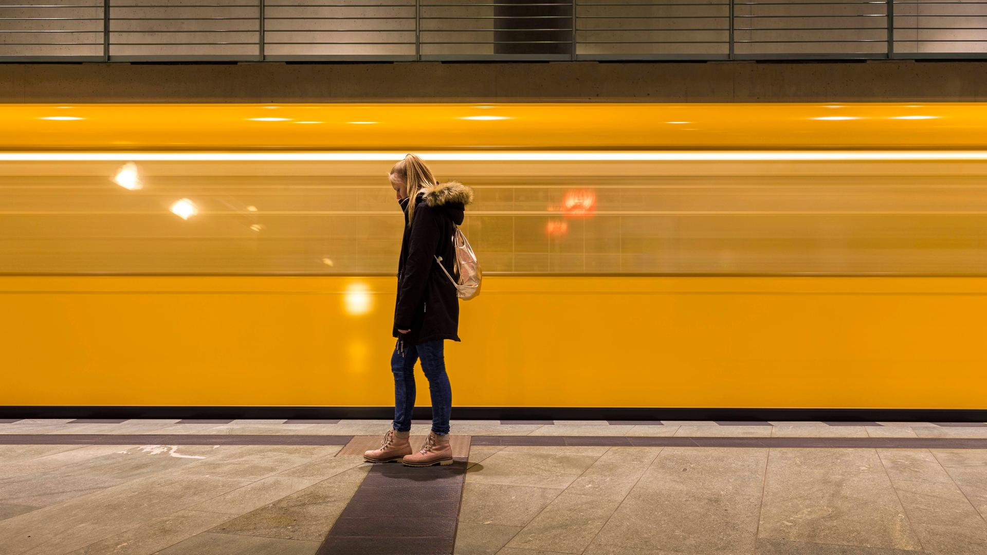 Eine junge Frau steht in einem U-Bahn-Schacht. Im Hintergrund fährt eine gelbe U-Bahn vorbei. Sie ist aber nur verschommen zu sehen. 