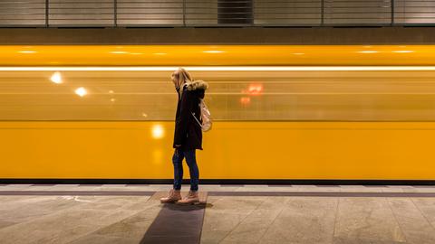 Eine junge Frau steht in einem U-Bahn-Schacht. Im Hintergrund fährt eine gelbe U-Bahn vorbei. Sie ist aber nur verschommen zu sehen. 