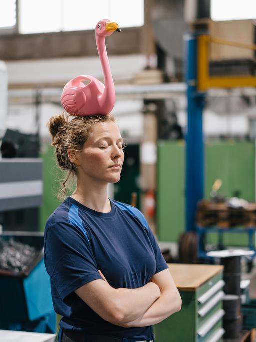 Eine Arbeiterin steht in einer Fertigungshalle mit verschränkten Armen und geschlossenen Augen. Auf ihrem Kopf balanciert ein pinker Plastikflamingo.