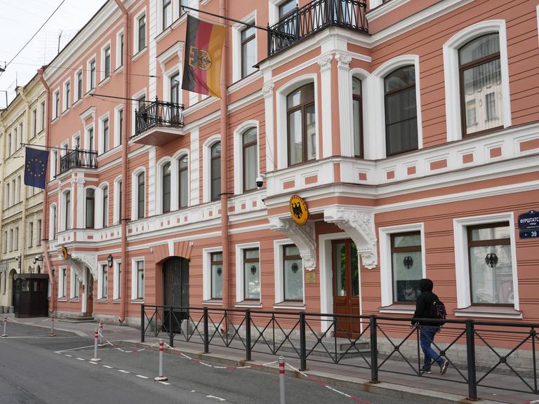 Das Bild zeigt ein Gebäude in St. Petersburg mit einer rot-weißen Fassade. Über der Tür hängen eine deutsche Fahne und ein deutsche Wappen.