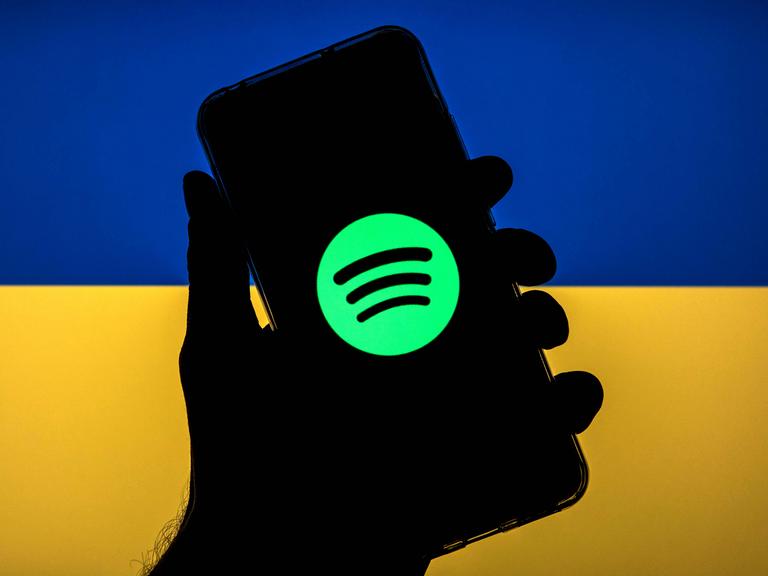 Hinter einem hochgehaltenen Handy, auf dem ein Streamingdienst angewählt wurde, leuchten die Farben der Ukraine: blau und gelb.