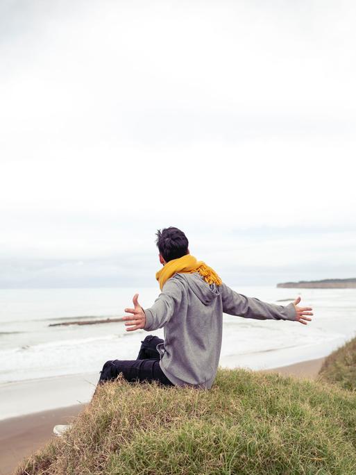 Ein Mensch sitzt am Meer und hat die Arme geöffnet. Ein Mensch sitzt am Meer und hat die Arme geöffnet.
