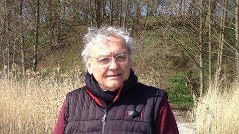 Ein älterer Mann mit Schal und Weste steht im Freien vor einem Hügel