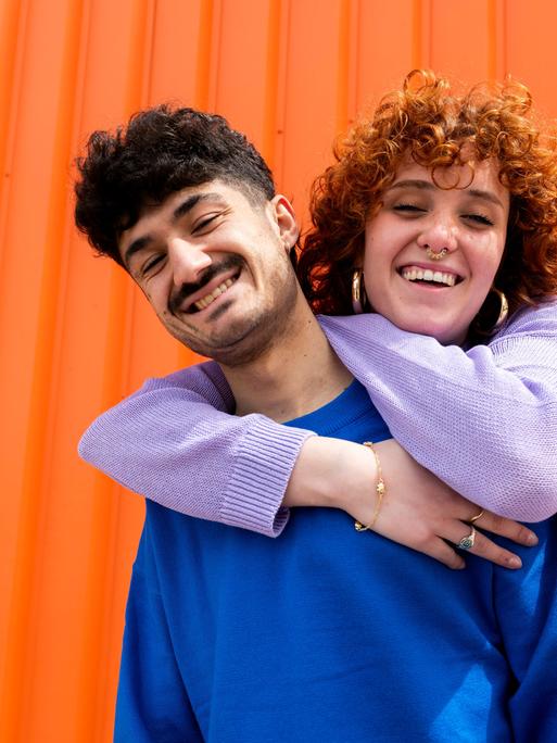 Eine Frau umarmt einen Freund von hinten. Im Hintergrund ein orangefarbener Container.