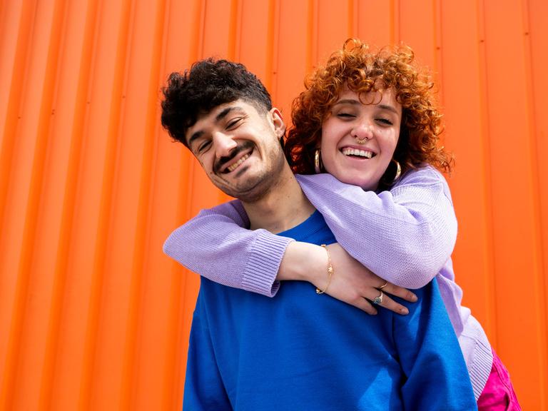 Eine Frau umarmt einen Freund von hinten. Im Hintergrund ein orangefarbener Container.