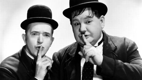 Stan Laurel und Oliver Hardy halten ihre Finger an den Mund als Zeichen für das Schweigen.