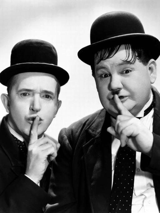 Stan Laurel und Oliver Hardy halten ihre Finger an den Mund als Zeichen für das Schweigen.