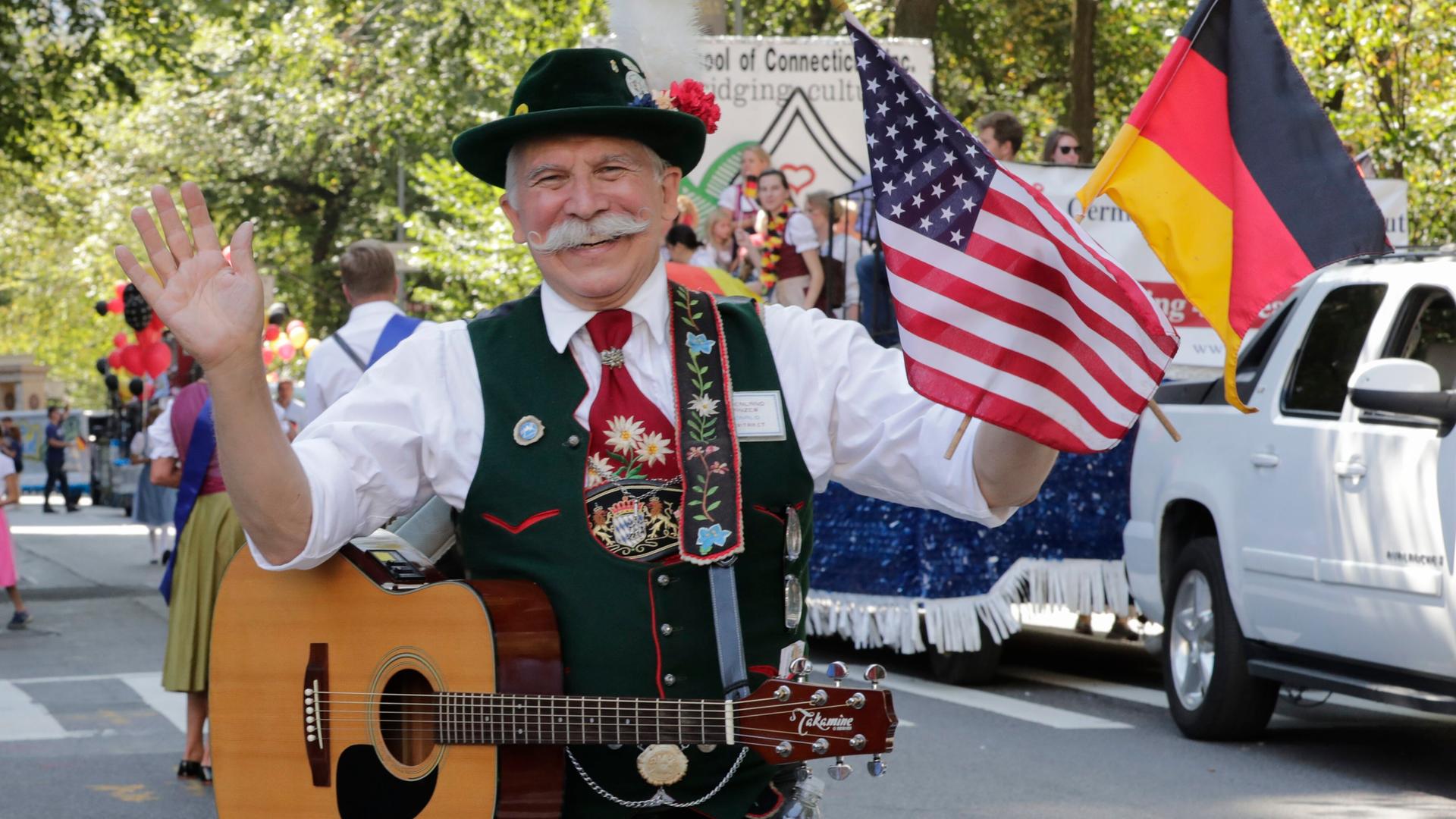 Ein älterer Mann in bestickter Tracht und mit Schnurrbart hält eine deutsche und eine US-amerikanische Flagge und lacht in die Kamera.