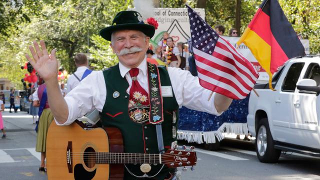 Ein älterer Mann in bestickter Tracht und mit Schnurrbart hält eine deutsche und eine US-amerikanische Flagge und lacht in die Kamera.