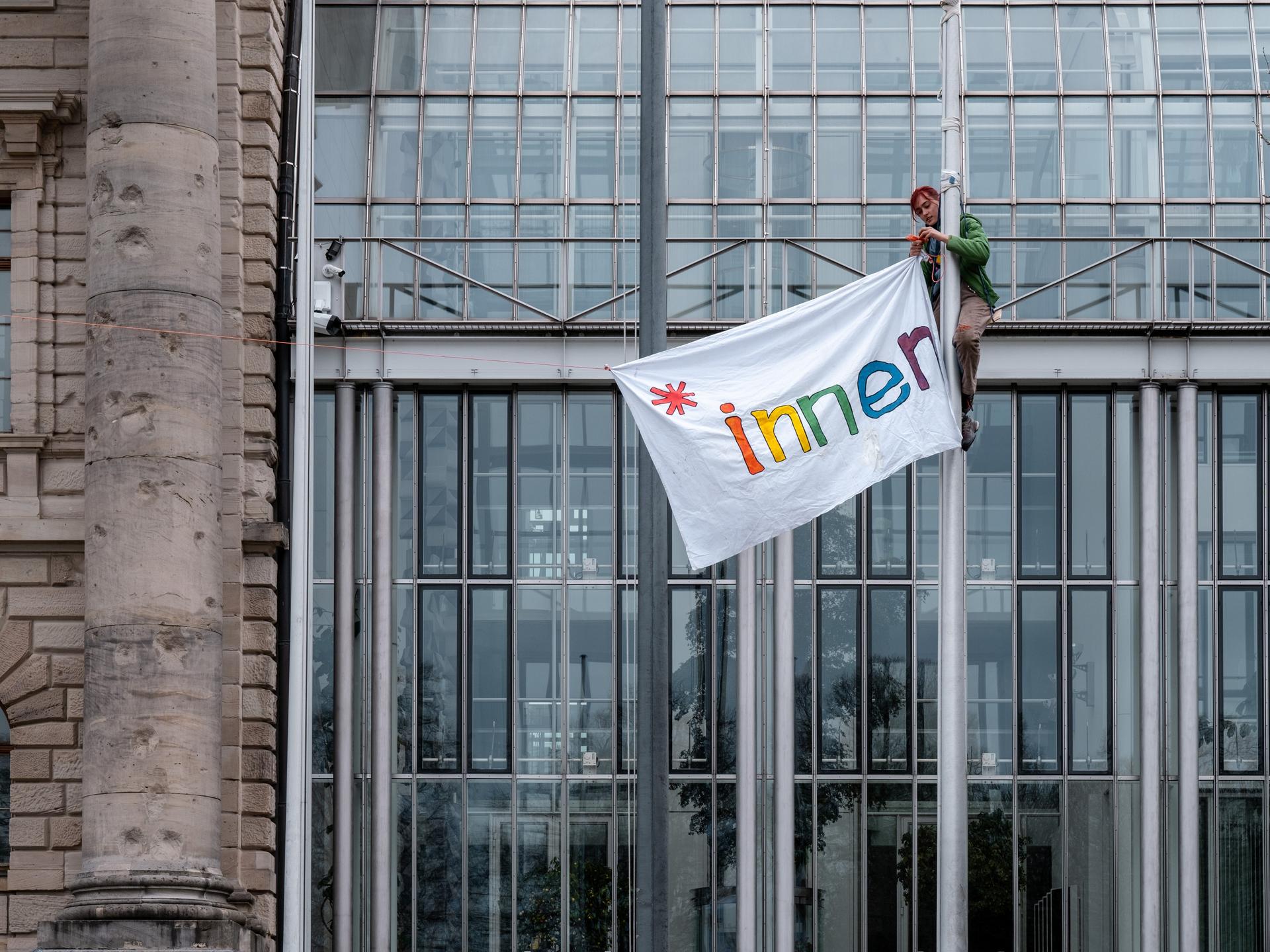 Vor der Bayerischen Staatskanzlei in München hissen Protestierende an Fahnenmasten ein Banner mit der Aufschift "*innen".