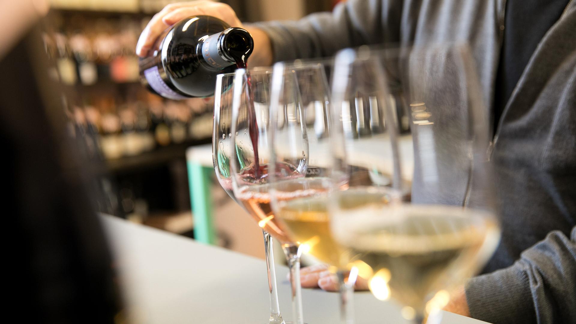 Mehrere Weingläser stehen in einer Reihe, in eins davon schenkt eine Person einen Schluck Rotwein ein.