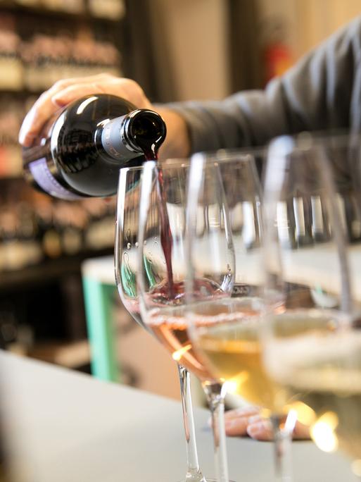 Mehrere Weingläser stehen in einer Reihe, in eins davon schenkt eine Person einen Schluck Rotwein ein.