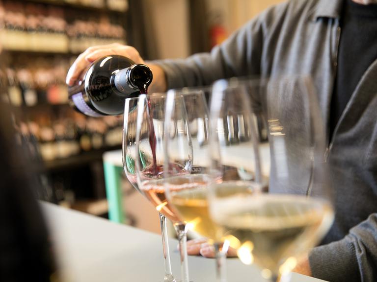 Mehrere Weingläser stehen in einer Reihe, in eins davon schenkt eine Person einen Schluck Rotwein ein.