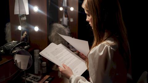 Eine junge Frau sitzt vor einem Schminkspiegel hinter der Bühne und schaut konzentriert auf Blätter eines Textheftes.