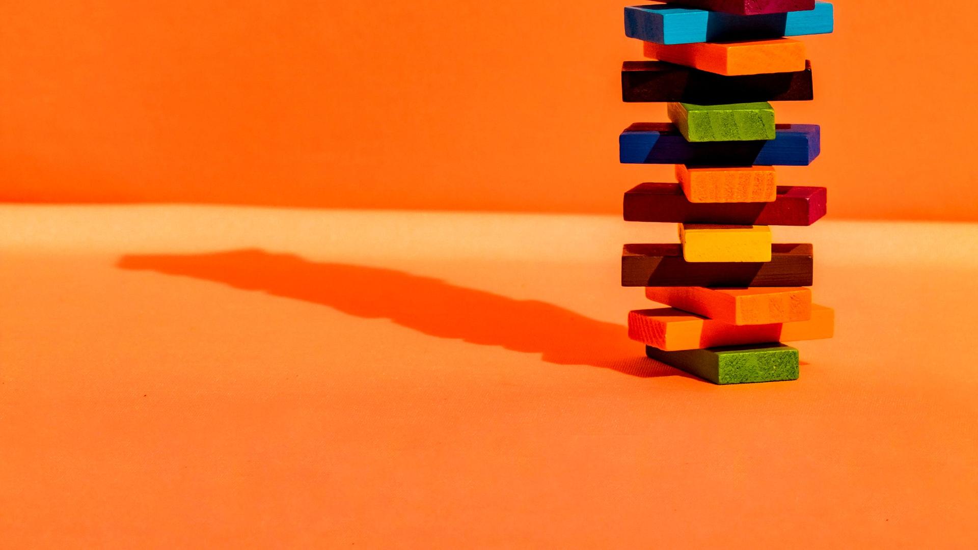 Ein Stapelturm aus bunten Bauklötzen vor orangefarbenem Hintergrund. Der Turm wirft einen kleinen Schatten auf der linken Seite.