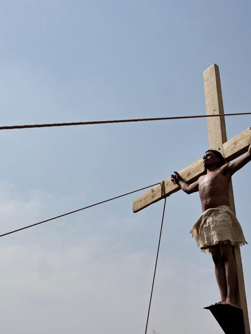 Ein Darsteller steht an einem Holzkreuz und imitiert die Kreuzigung Jesu. Im Hintergrund blauer Himmel.