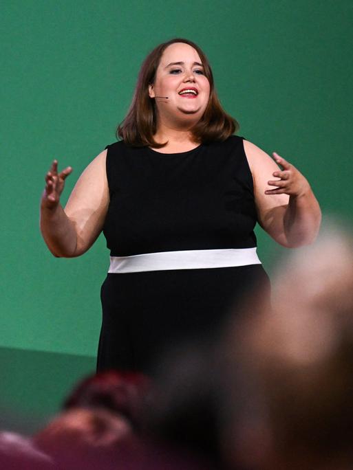 Ricarda Lang steht im schwarzen Kleid auf einer Bühne und spricht zu Publikum.