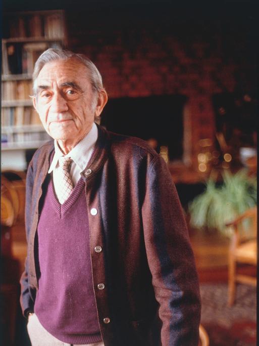 Pierre Schaeffer in Porträtaufnahme in seiner Wohnung, in der er vor Bücherregalen steht.