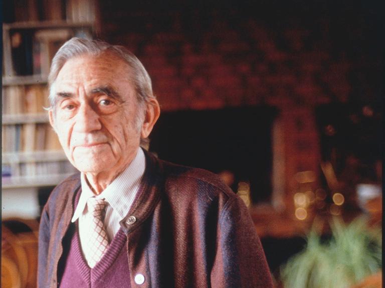 Pierre Schaeffer in Porträtaufnahme in seiner Wohnung, in der er vor Bücherregalen steht.