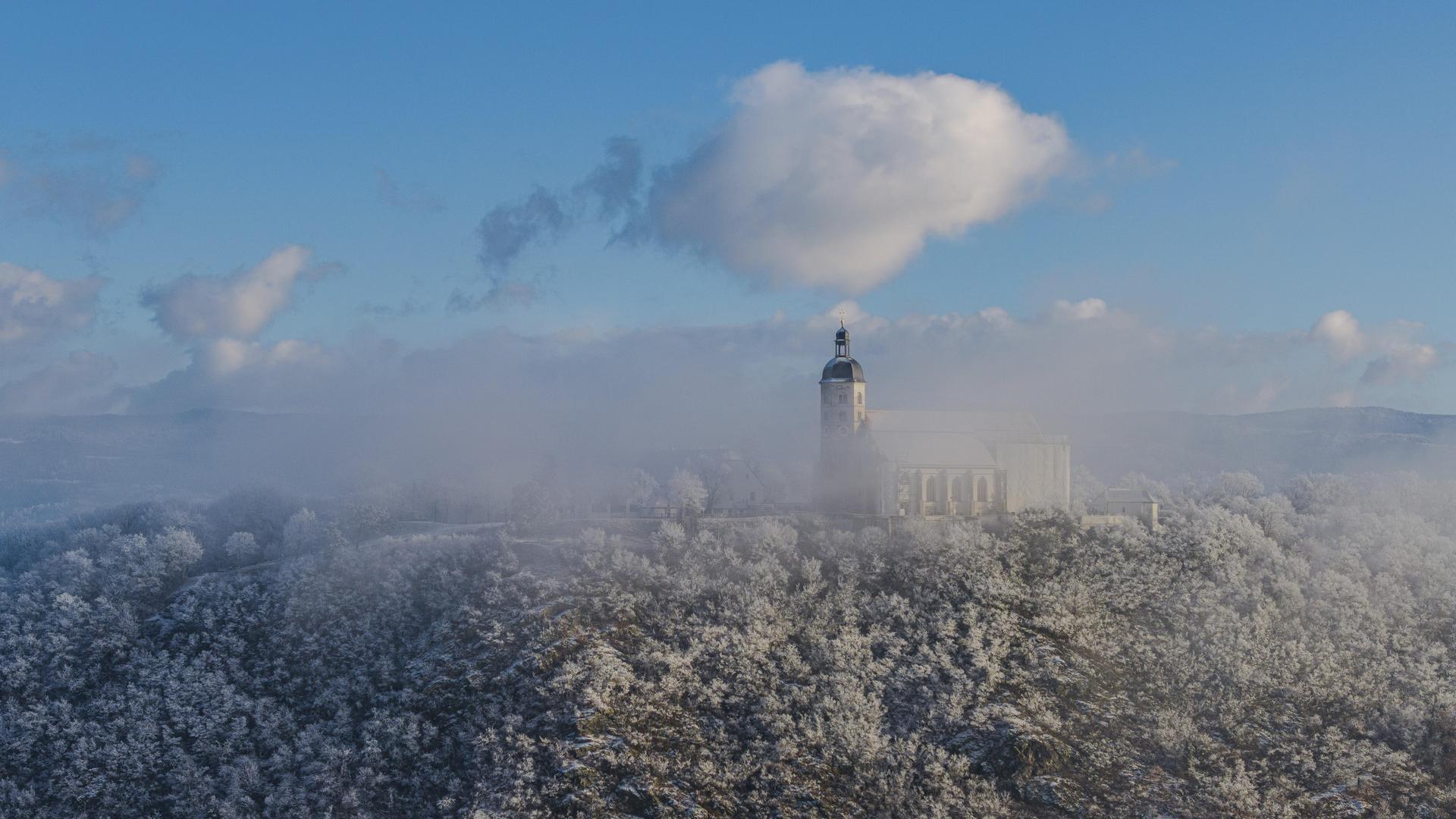 Die Szene zeigt eine winterliche Landschaft mit schneebedeckten Bäumen und der Wallfahrtskirche Mariä Himmelfahrt auf dem Bogenberg. Der Himmel hat einige Wolken und das Licht ist klar. 