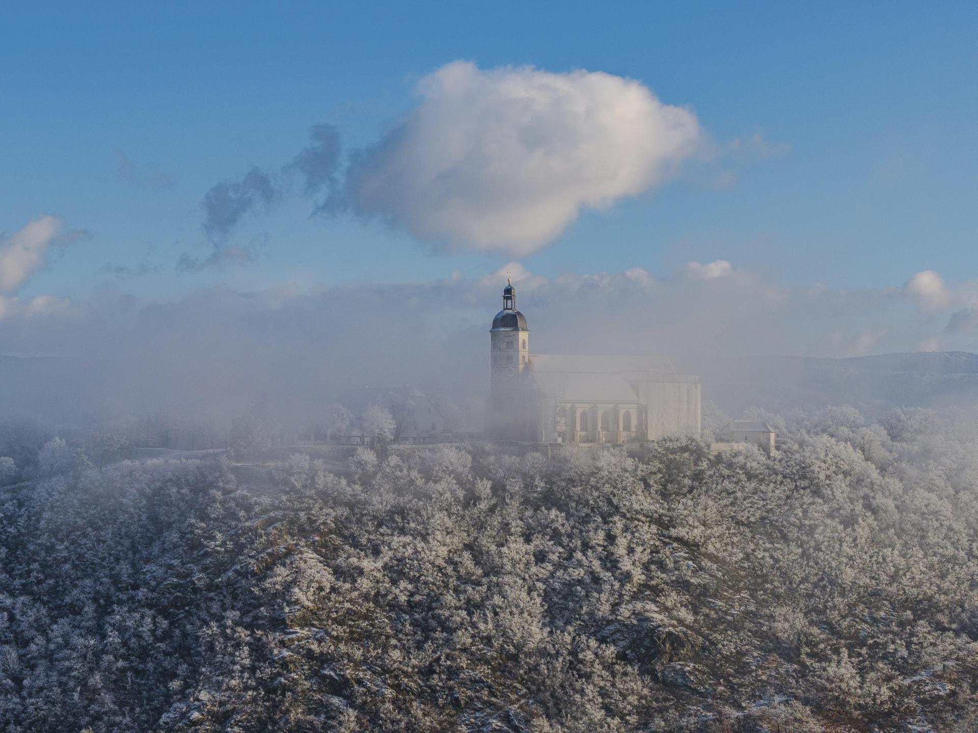 Die Szene zeigt eine winterliche Landschaft mit schneebedeckten Bäumen und der Wallfahrtskirche Mariä Himmelfahrt auf dem Bogenberg. Der Himmel hat einige Wolken und das Licht ist klar. 