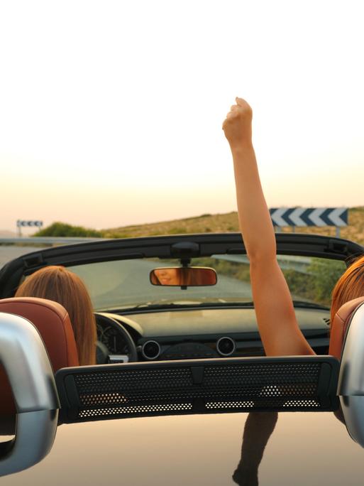 Zwei Frauen in einem Cabriolet. Eine reckt die Hände in die Luft.