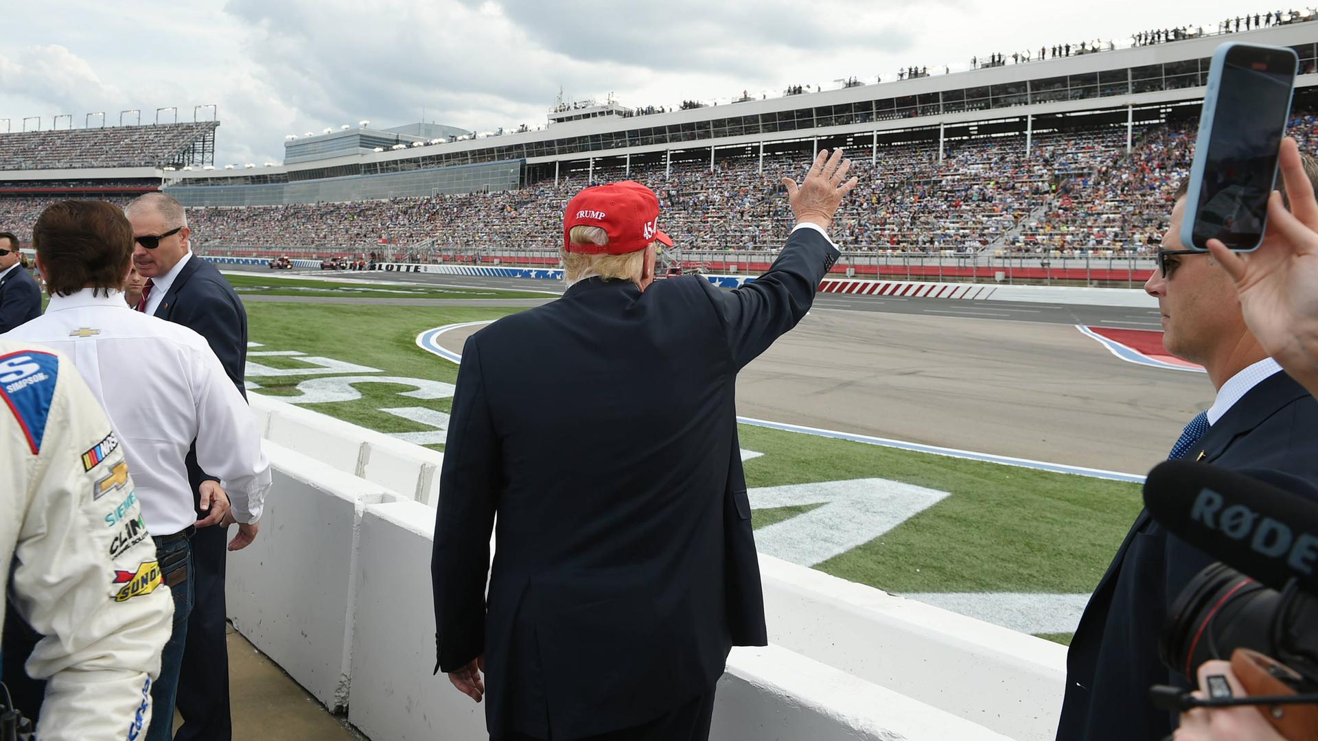 Donald Trump steht mit dem Rücken zur Kamera. Er winkt in Richtung einer sehr großen Tribüne voller Menschen auf der anderen Seite der Rennstrecke.