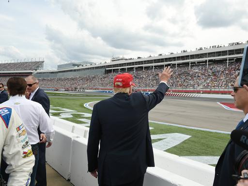 Donald Trump steht mit dem Rücken zur Kamera. Er winkt in Richtung einer sehr großen Tribüne voller Menschen auf der anderen Seite der Rennstrecke.