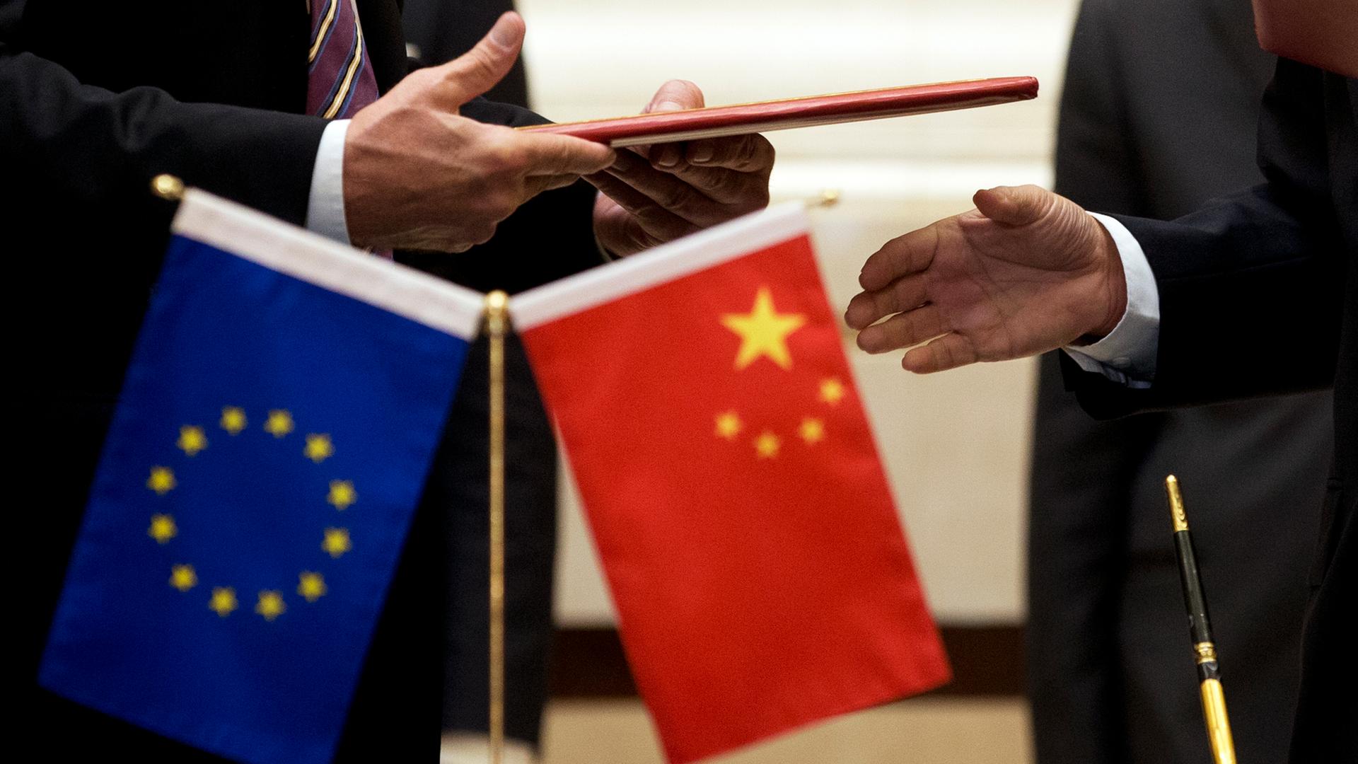 Im Vordergrund sind die beiden Flaggen von China und der Europäischen Union zu sehen. Diese stehen auf einem Tisch. Im Hintergrund stehen zwei Menschen. Ihre Hände sind zu sehen. 