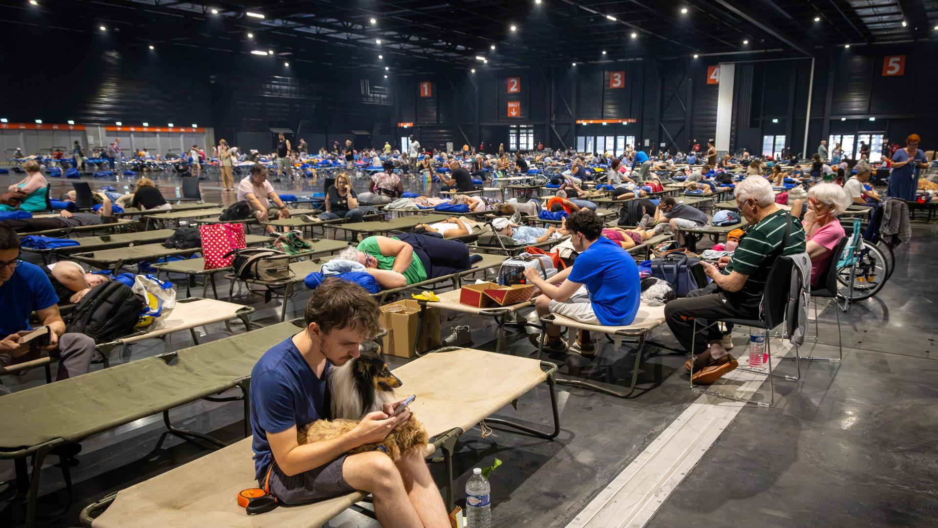 Das Foto zeigt eine Halle in Bordeaux, in der viele Menschen auf Feldbetten sitzen und liegen.