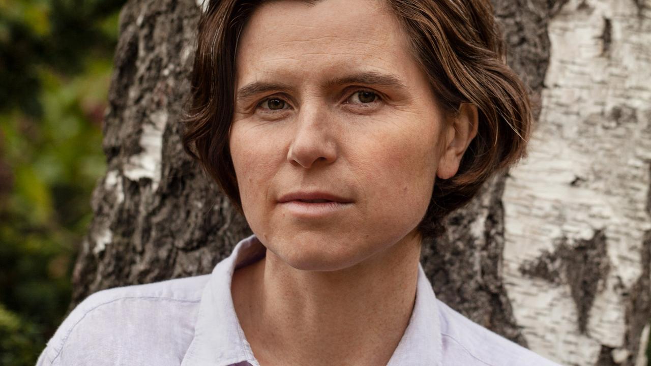 Eine Frau mit kurzem braunen Haar blickt in die Kamera. Sie steht vor einer Birke und trägt ein helles Hemd mit Kragen.