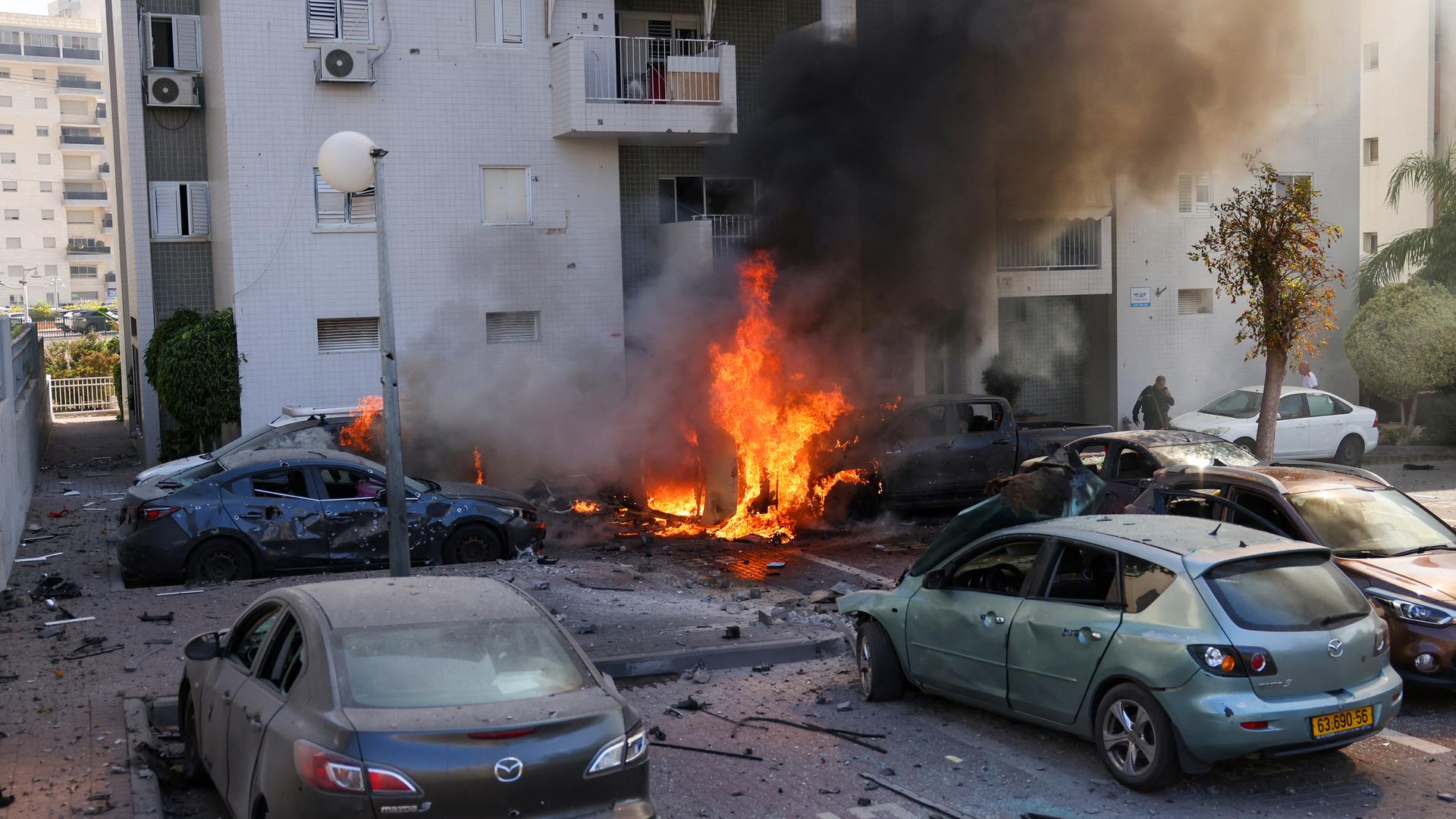 Feuer vor einem Wohnhaus, ein Mitglied der israelischen Sicherheitskräfte steht in der Nähe brennender Autos nach einem Raketenangriff aus dem Gazastreifen in Aschkelon