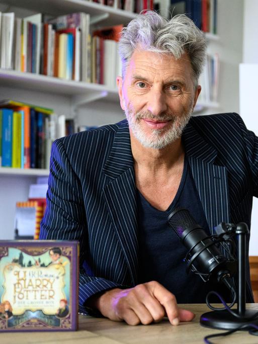 Der Schauspieler Rufus Beck sitzt an einem Tisch vor einer Bücherwand, links steht eine CD mit der Aufschrift "Harry Potter", rechts hinter Beck ist eine Zeichnung zu sehen, die Harry Potter darstellt, ein Kind mit einer Brille.